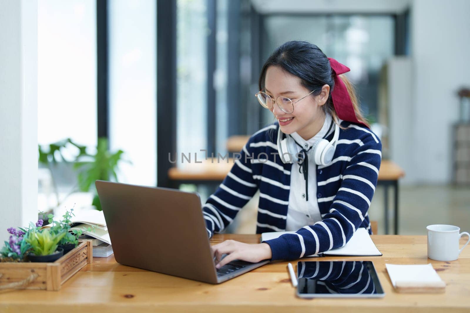 Portrait of a teenage Asian woman using a computer, wearing headphones and using a notebook to study online via video conferencing on a wooden desk in library.