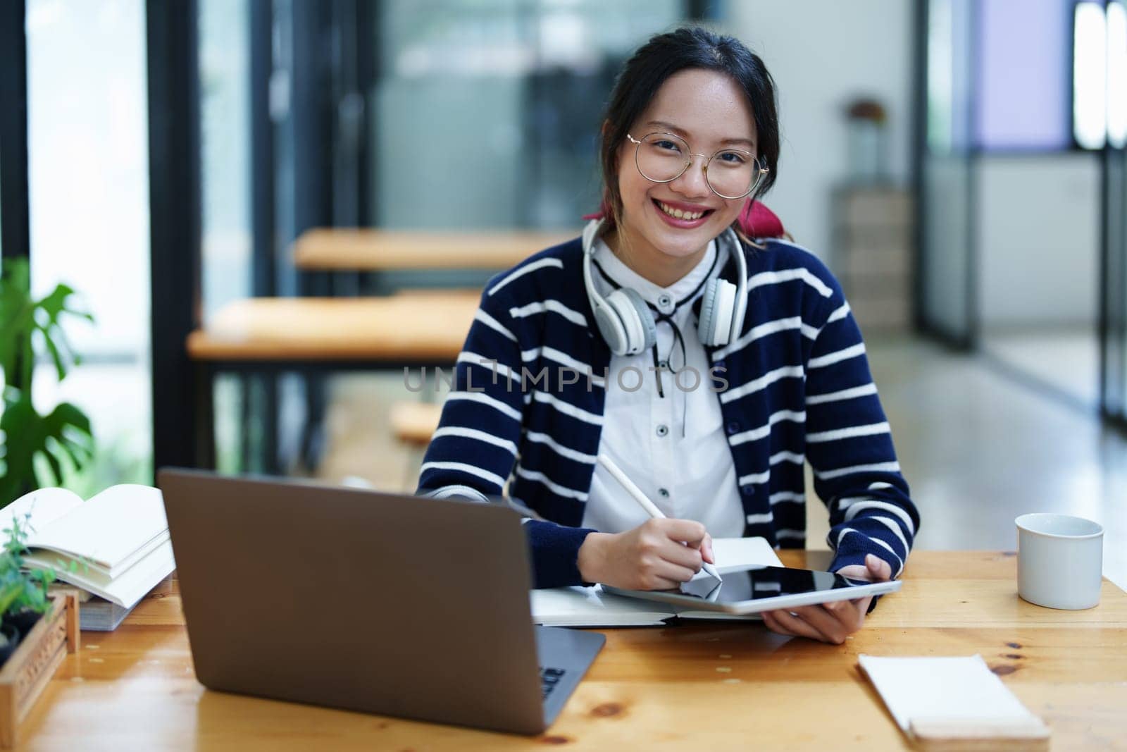 Portrait of a teenage Asian woman using a tablet, wearing headphones and using a notebook to study online via video conferencing on a wooden desk in library.