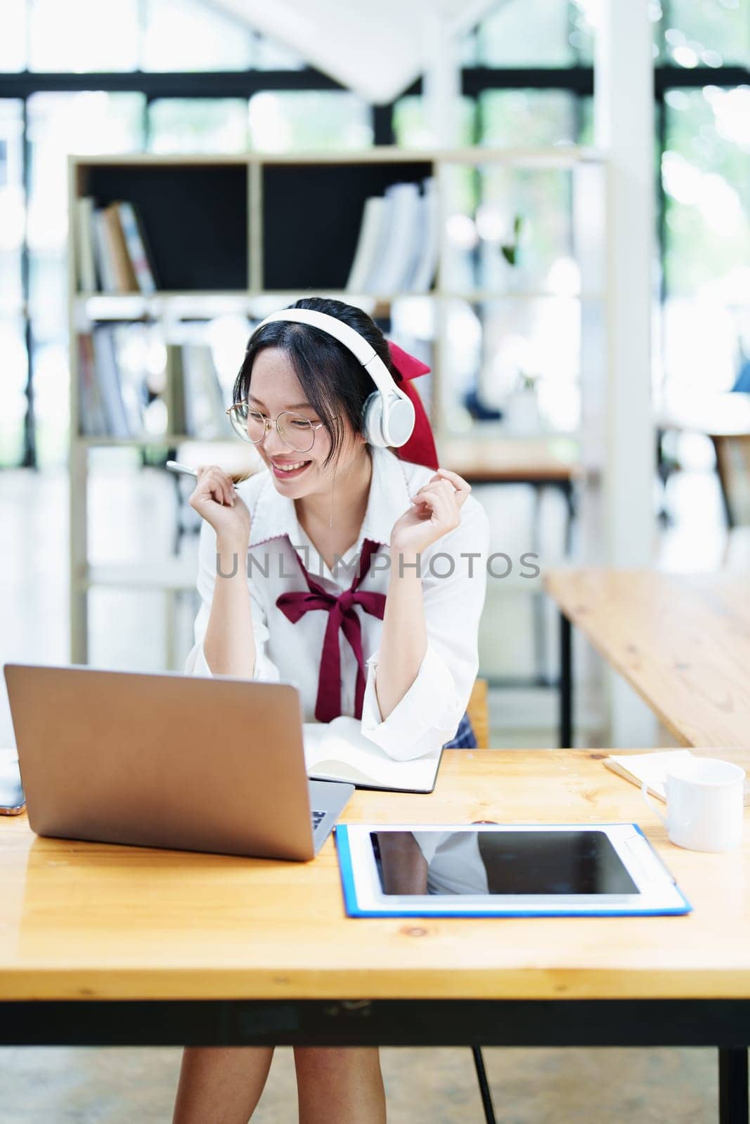 Portrait of a teenage Asian woman using a computer, wearing headphones and using a notebook to study online via video conferencing on a wooden desk in library by Manastrong