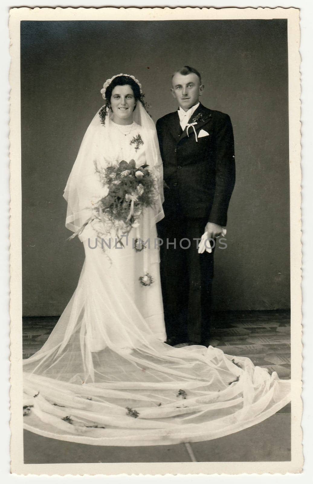 Vintage photo of newlyweds. Bride wears veil, long wedding gown and holds wedding bouquet. Groom wears black suit and white bow tie. Black white antique studio portrait. by roman_nerud