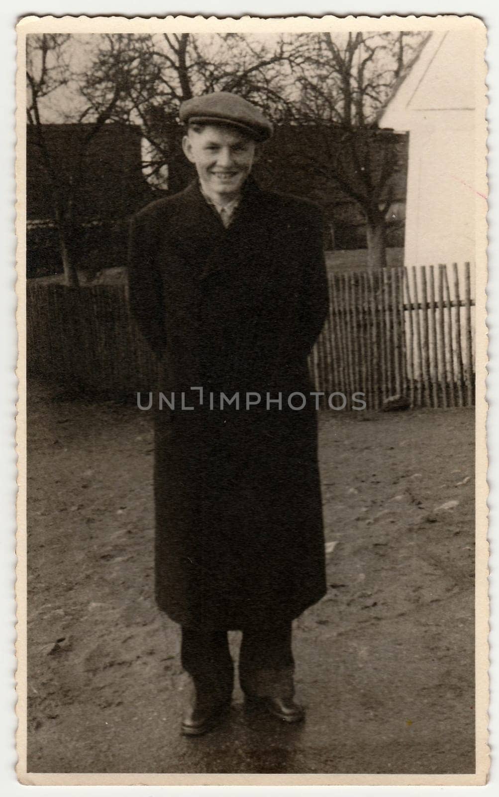 Vintage photo shows young man wears peaked cap, poses outdoors. Wooden fence and house is on background. Black white photography. by roman_nerud