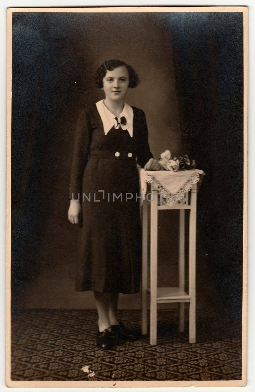 Vintage photo shows woman wears an elegant dress, poses next to white table. Black white antique studio photography with sepia effect. by roman_nerud