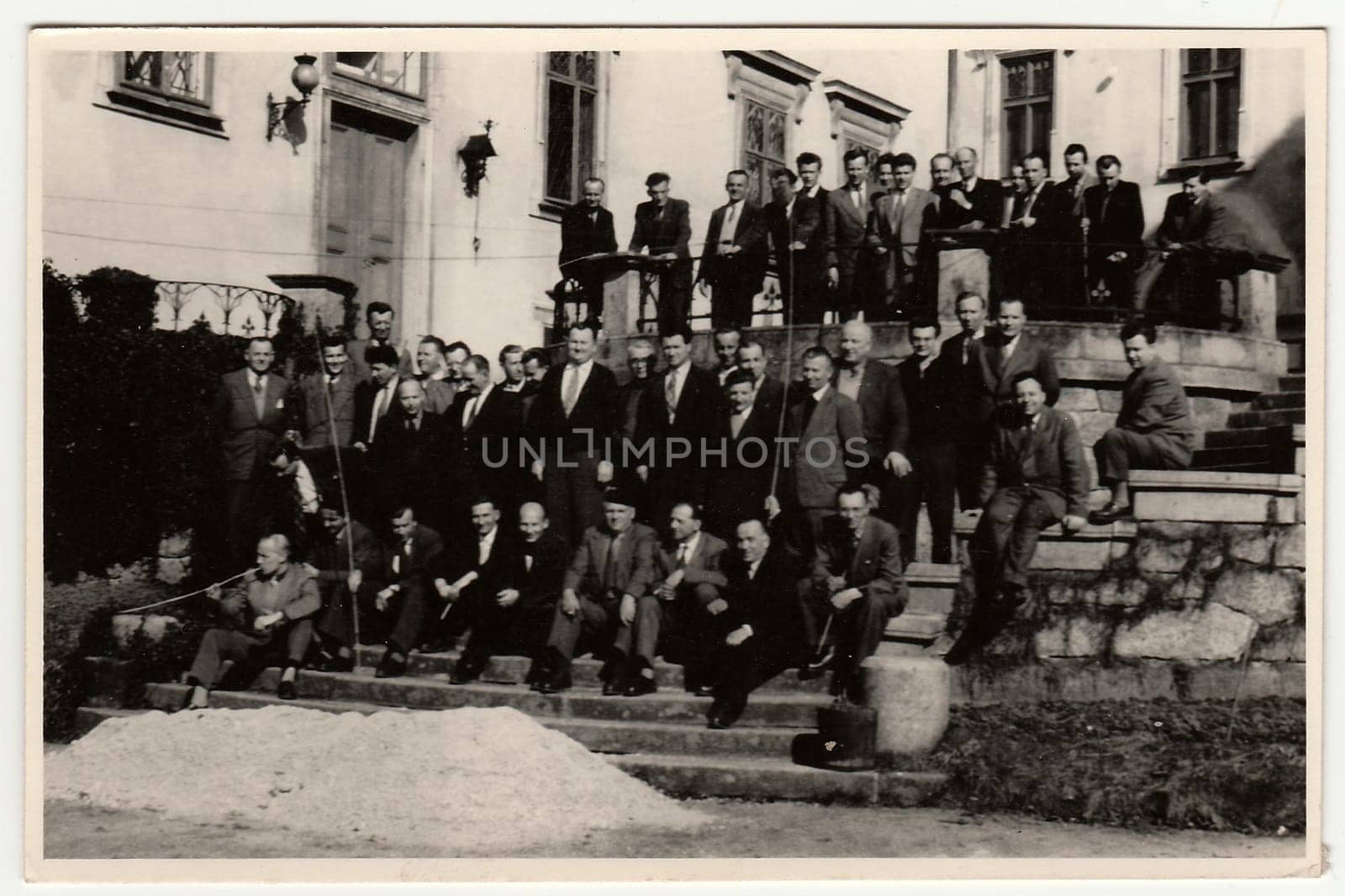 Vintage photo shows a big group of men stand in front of historic building. Antique black white photography. by roman_nerud