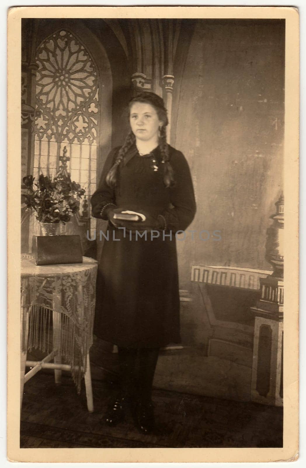 Vintage photo shows young woman poses with book, she stands next to the table with a bunch of flowers. Black white antique studio photography with sepia effect. by roman_nerud