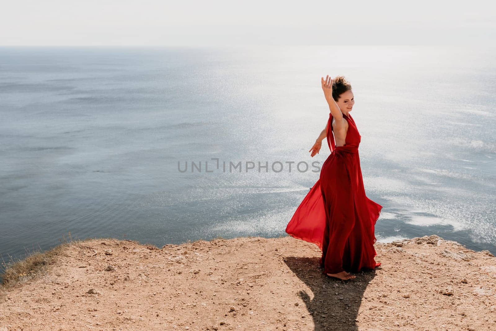 Side view a Young beautiful sensual woman in a red long dress posing on a rock high above the sea during sunrise. Girl on the nature on blue sky background. Fashion photo.