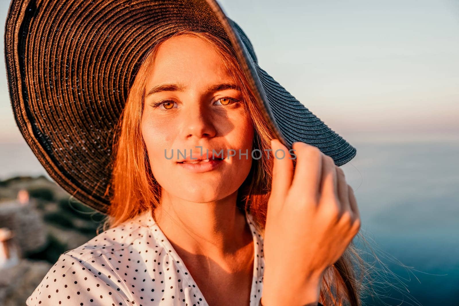 Portrait of happy young woman wearing summer black hat with large brim at beach on sunset. Closeup face of attractive girl with black straw hat. Happy young woman smiling and looking at camera at sea
