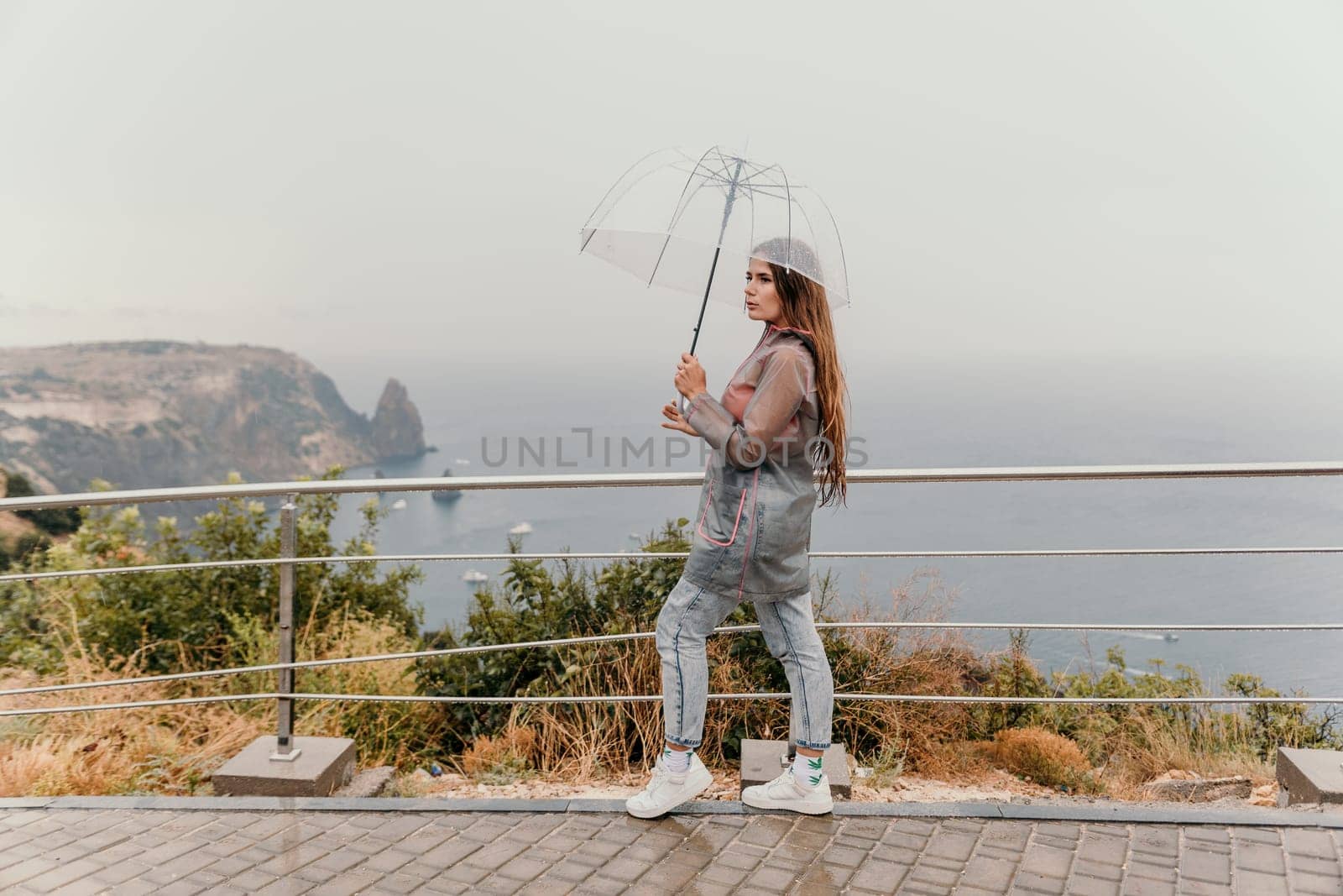 Woman rain park. Happy woman portrait wearing a raincoat with transparent umbrella outdoors on rainy day in park near sea. Girl on the nature on rainy overcast day