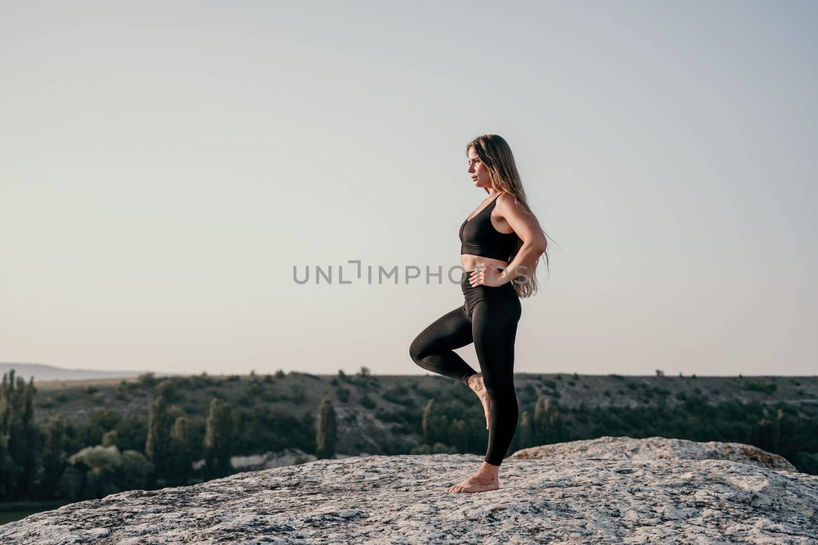 Well looking middle aged woman with long hair, fitness instructor in leggings and tops doing stretching and pilates on the rock near forest. Female fitness yoga routine concept. Healthy lifestyle.