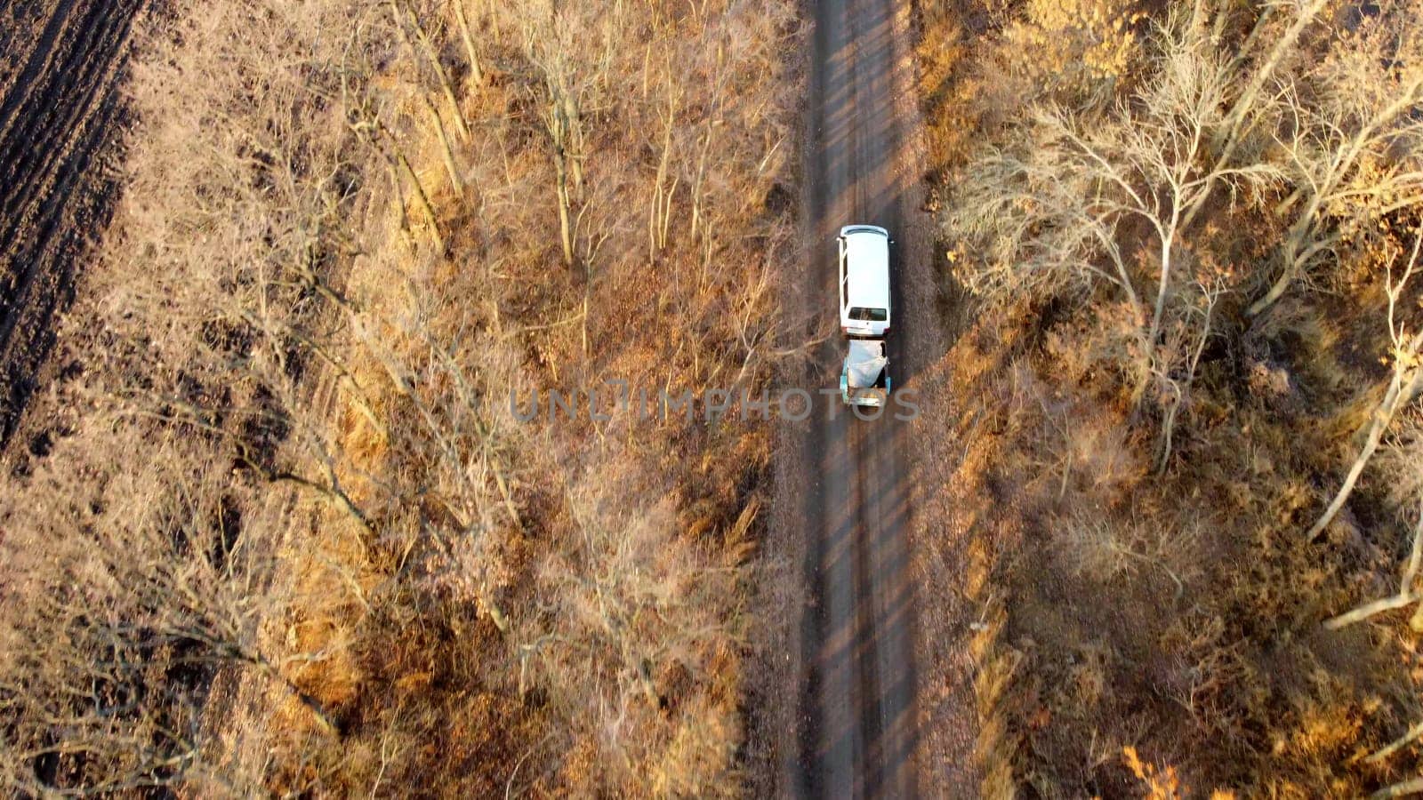 White car with trailer driving along dirt country road between trees without leaves on autumn sunny day. Rural countryside landscape, country scenery. Aerial drone view. Travel vehicle tourism