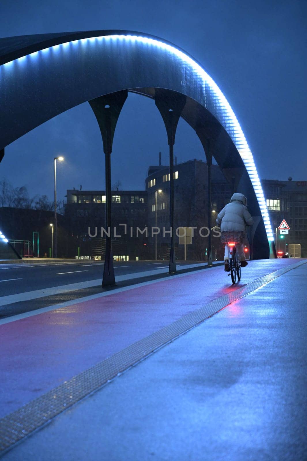 Woman with gray jacket riding a bicycle in rain over illuminated highway bridge in Heerdt