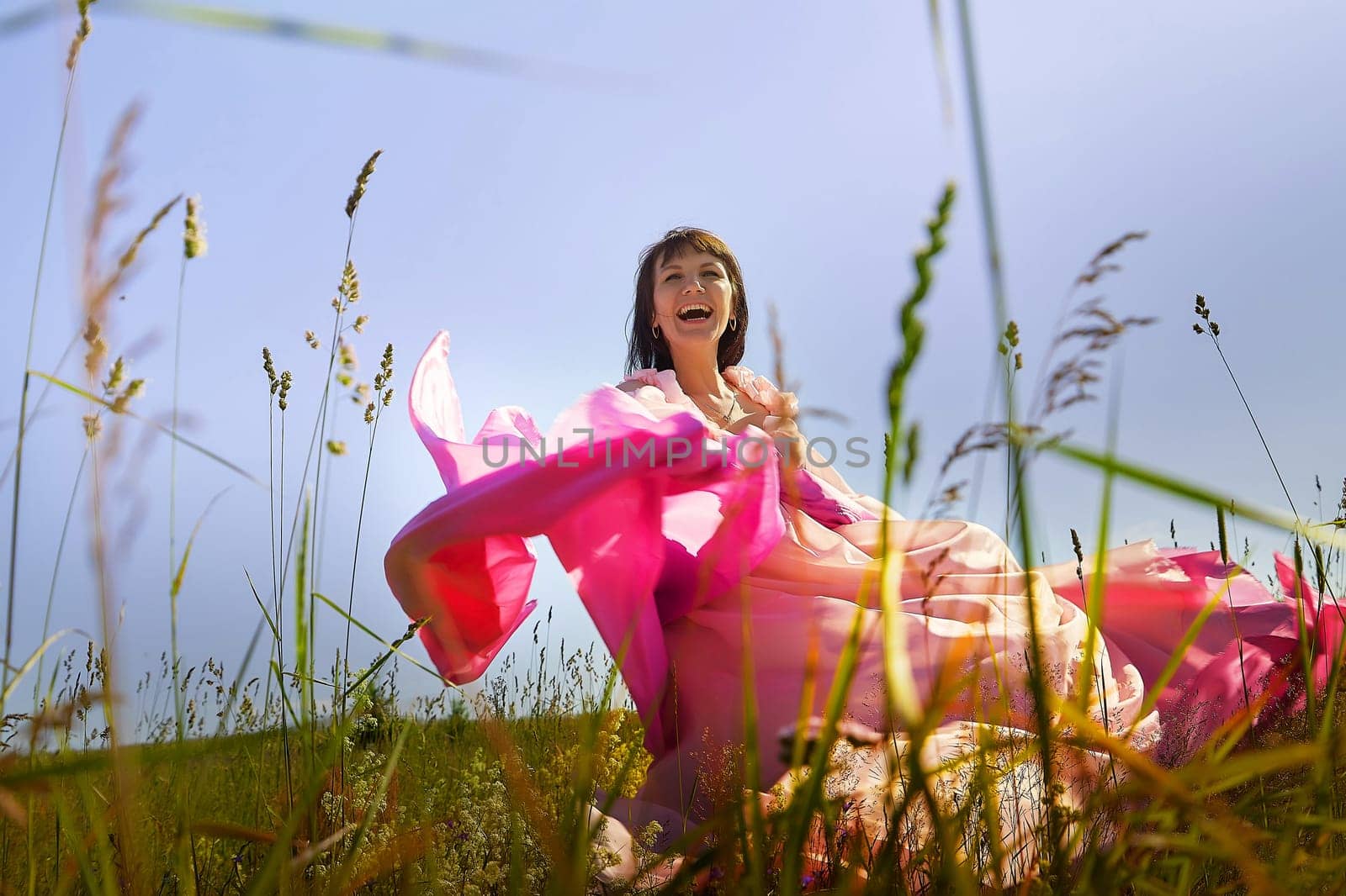 Beautiful girl in lush pink ball gown in green field during blooming of flowers and blue sky on background. Model posing on nature landscape as princess from fary tale