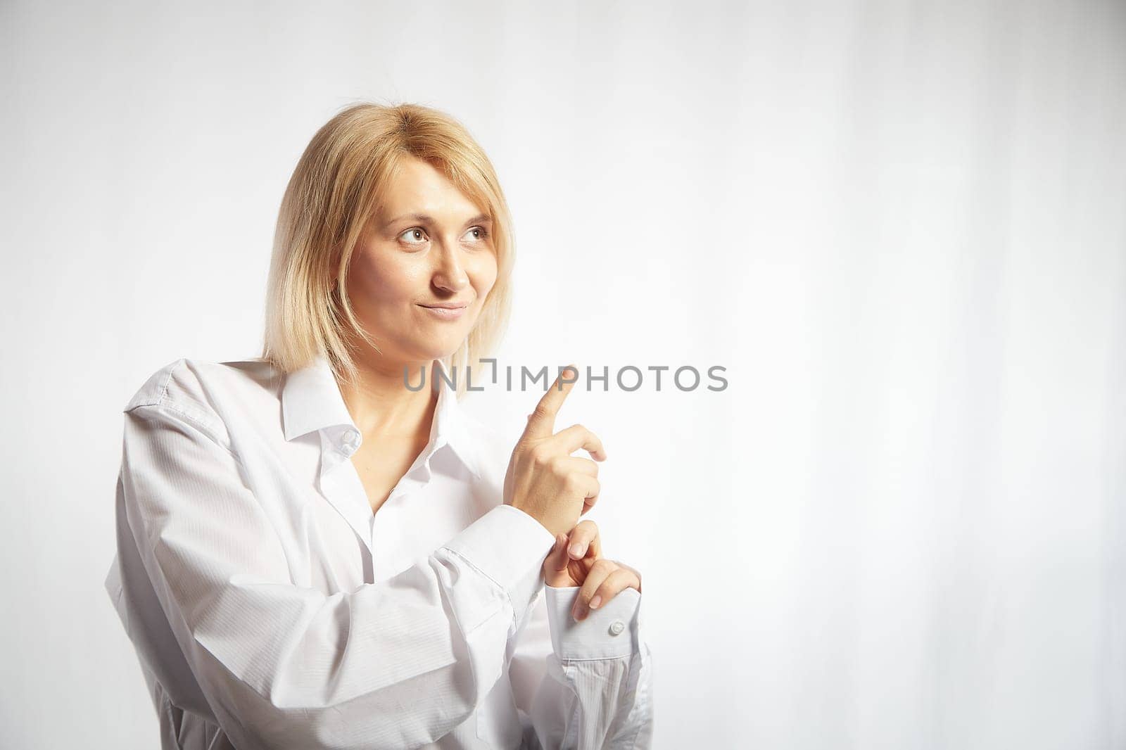 Portrait of a pretty blonde smiling woman posing on a white background and pointing somewhere. Happy girl model in white shirt in studio. Copy space by keleny