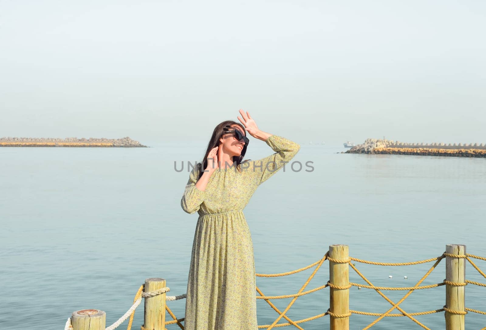 A young beautiful woman in a long dress stands alone on a pier by the sea, looking at the sun