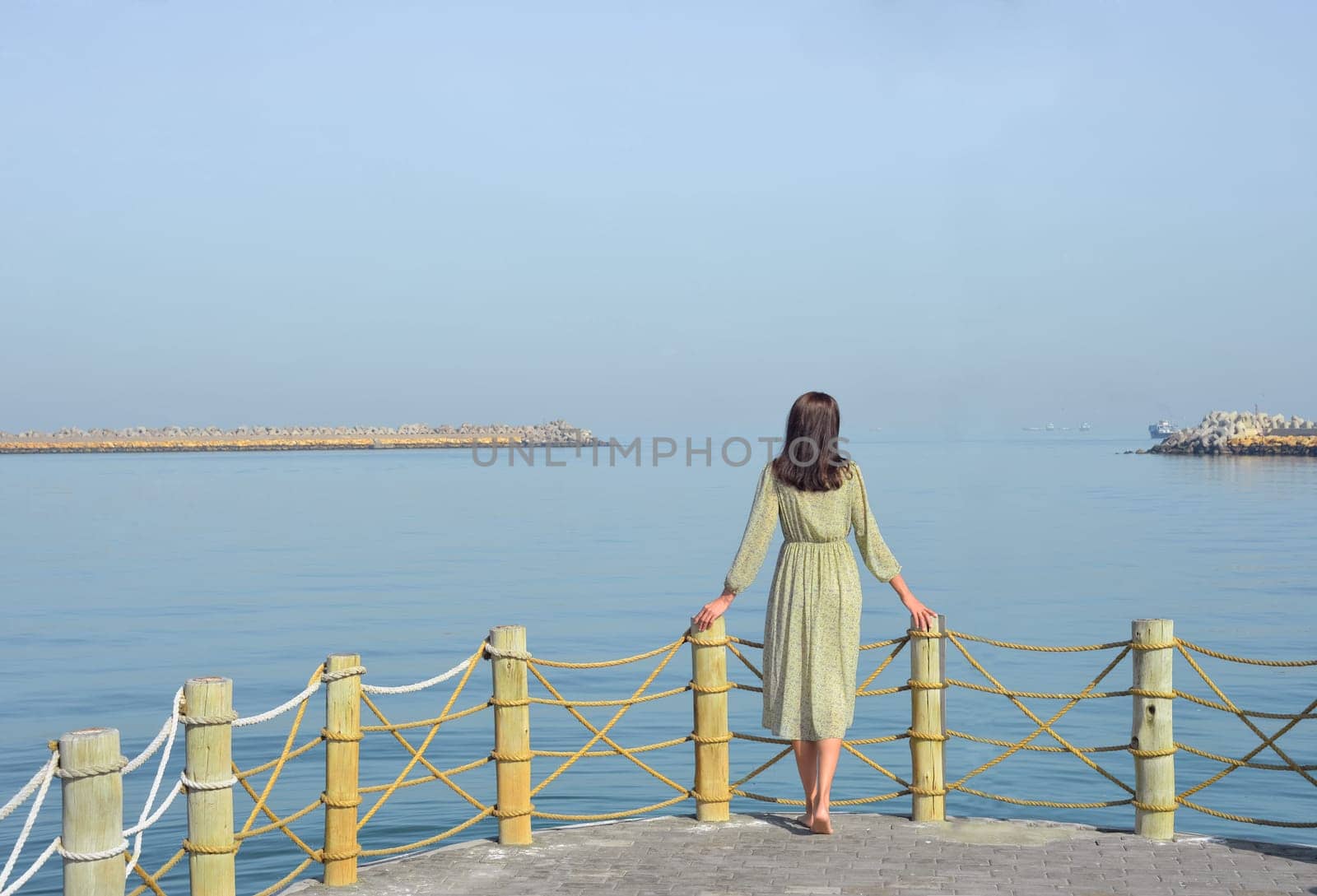 A young woman in a long dress with a backpack walks along the pier by the sea against the background of the developing UAE flag, and looks into the distance. by Ekaterina34