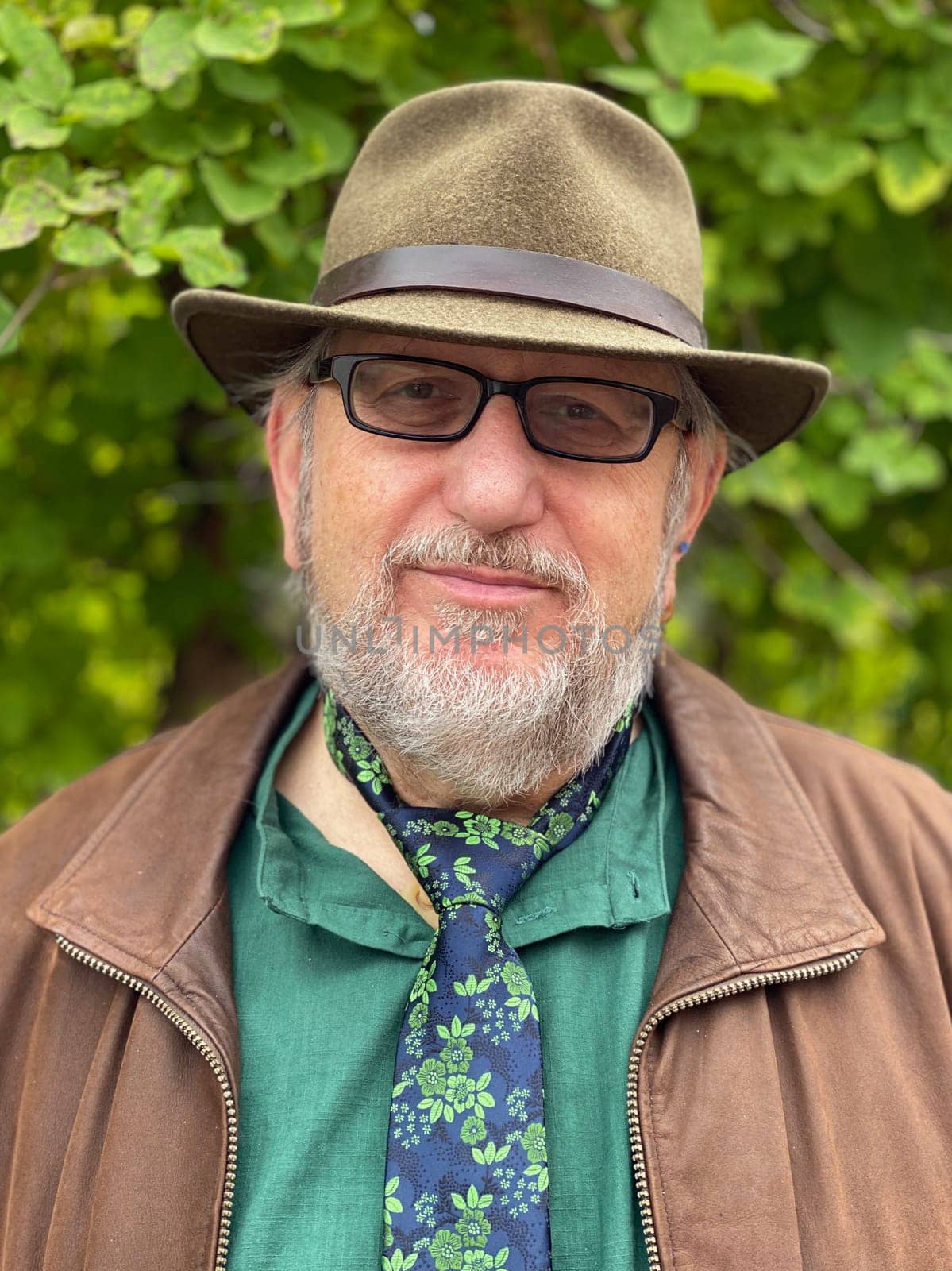 Elderly man with beard, glasses and hat looking into the camera. He wears his tie direct on his neck over his shirt. He's an individualist.