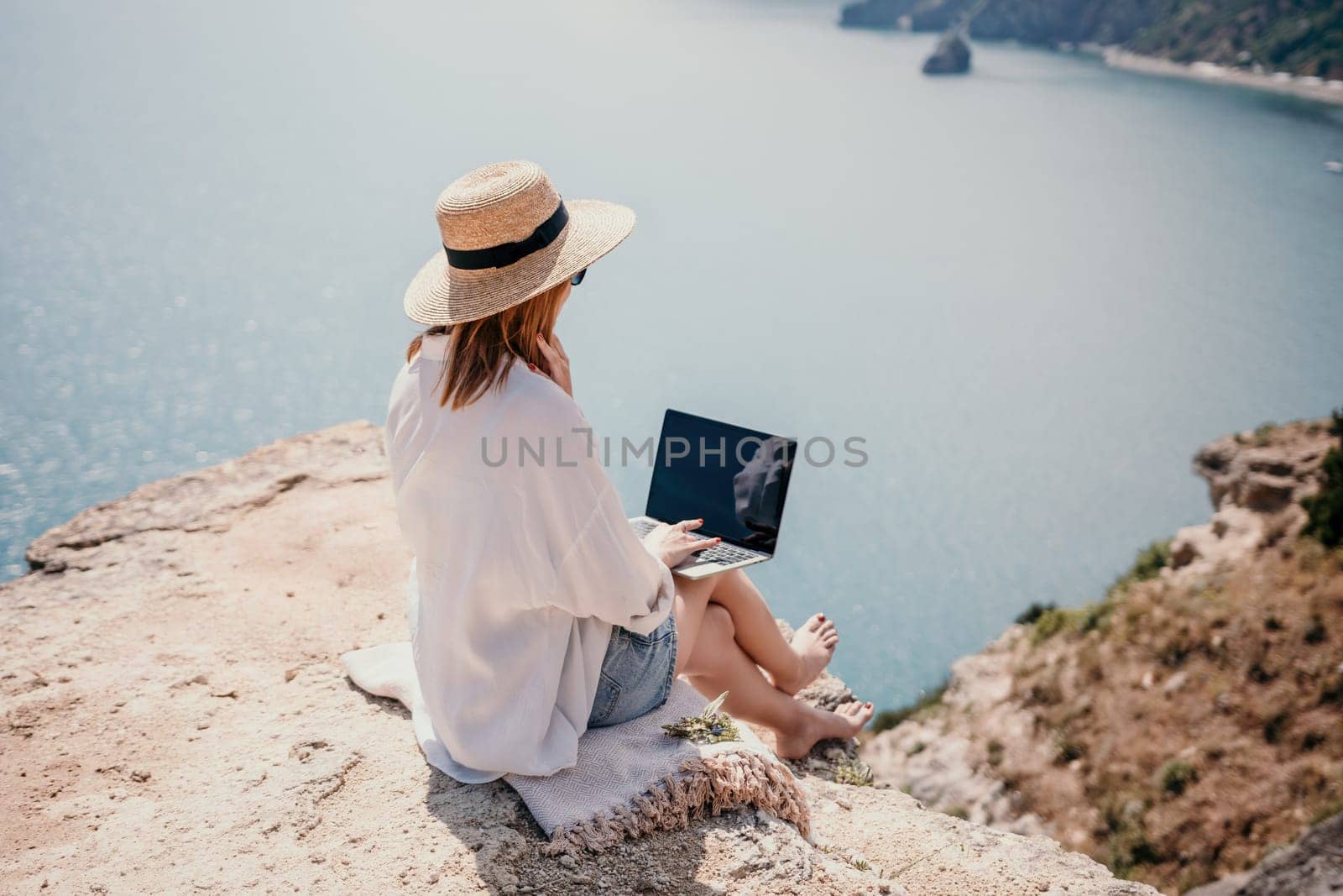 Successful business woman in yellow hat working on laptop by the sea. Pretty lady typing on computer at summer day outdoors. Freelance, travel and holidays concept.