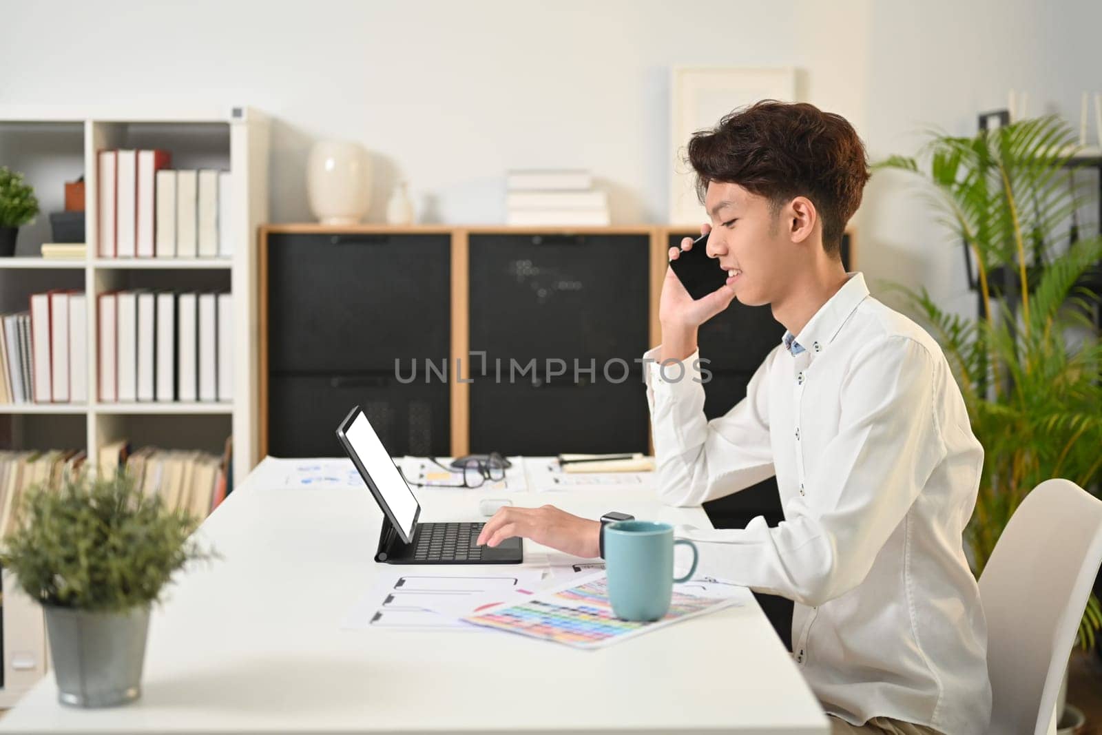 Side view of young creative man in white shirt using digital tablet and talking on mobile phone.