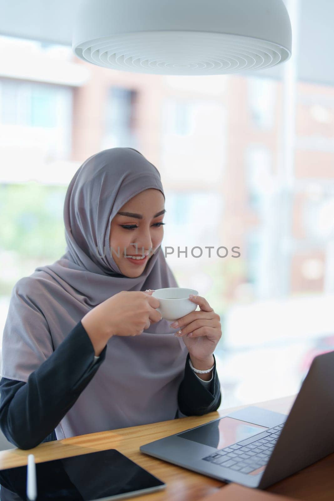A beautiful Muslim woman with a smiling face in the morning drinking coffee and using a computer.