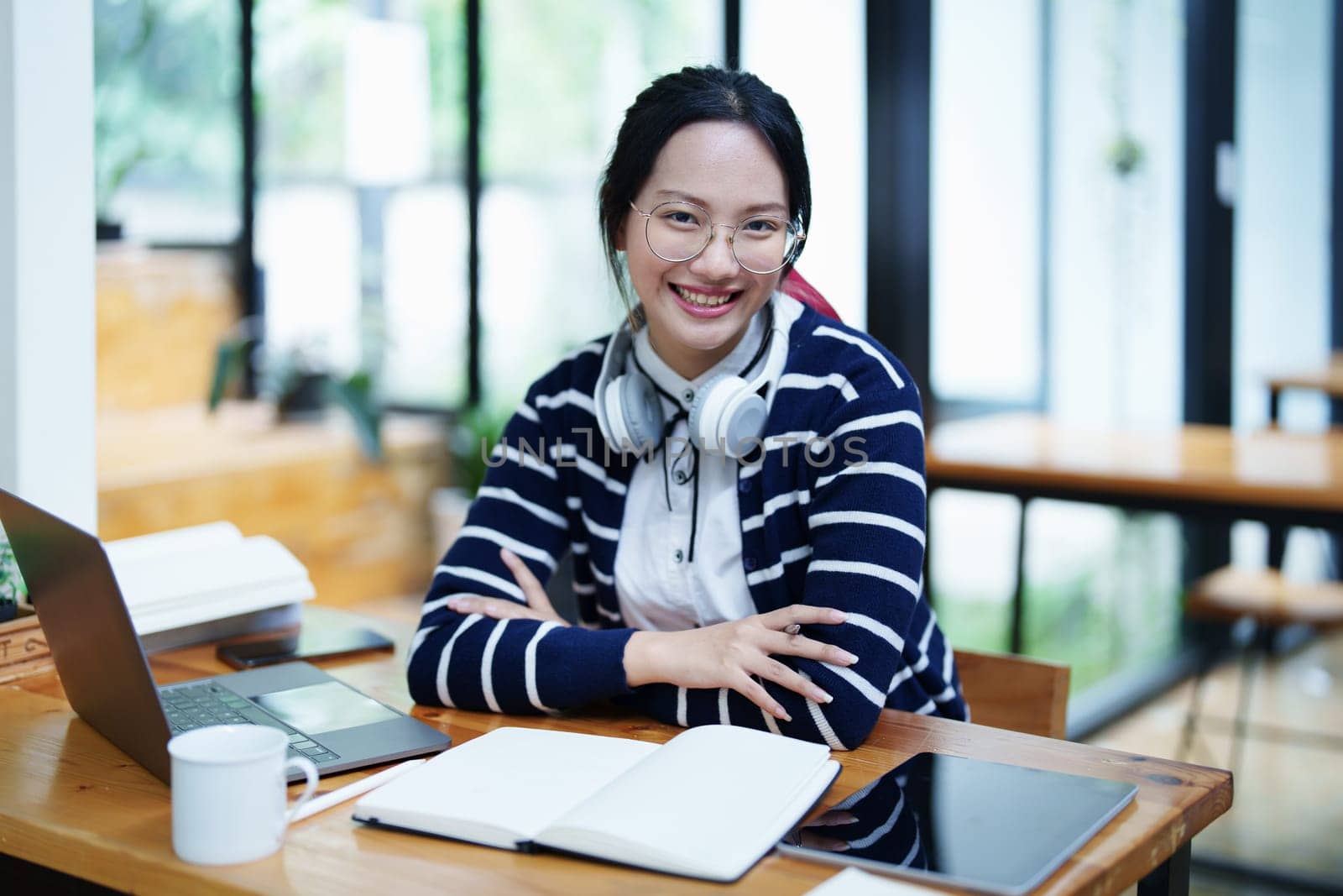 Portrait of a teenage Asian woman using a computer, wearing headphones and using a notebook to study online via video conferencing on a wooden desk in library by Manastrong