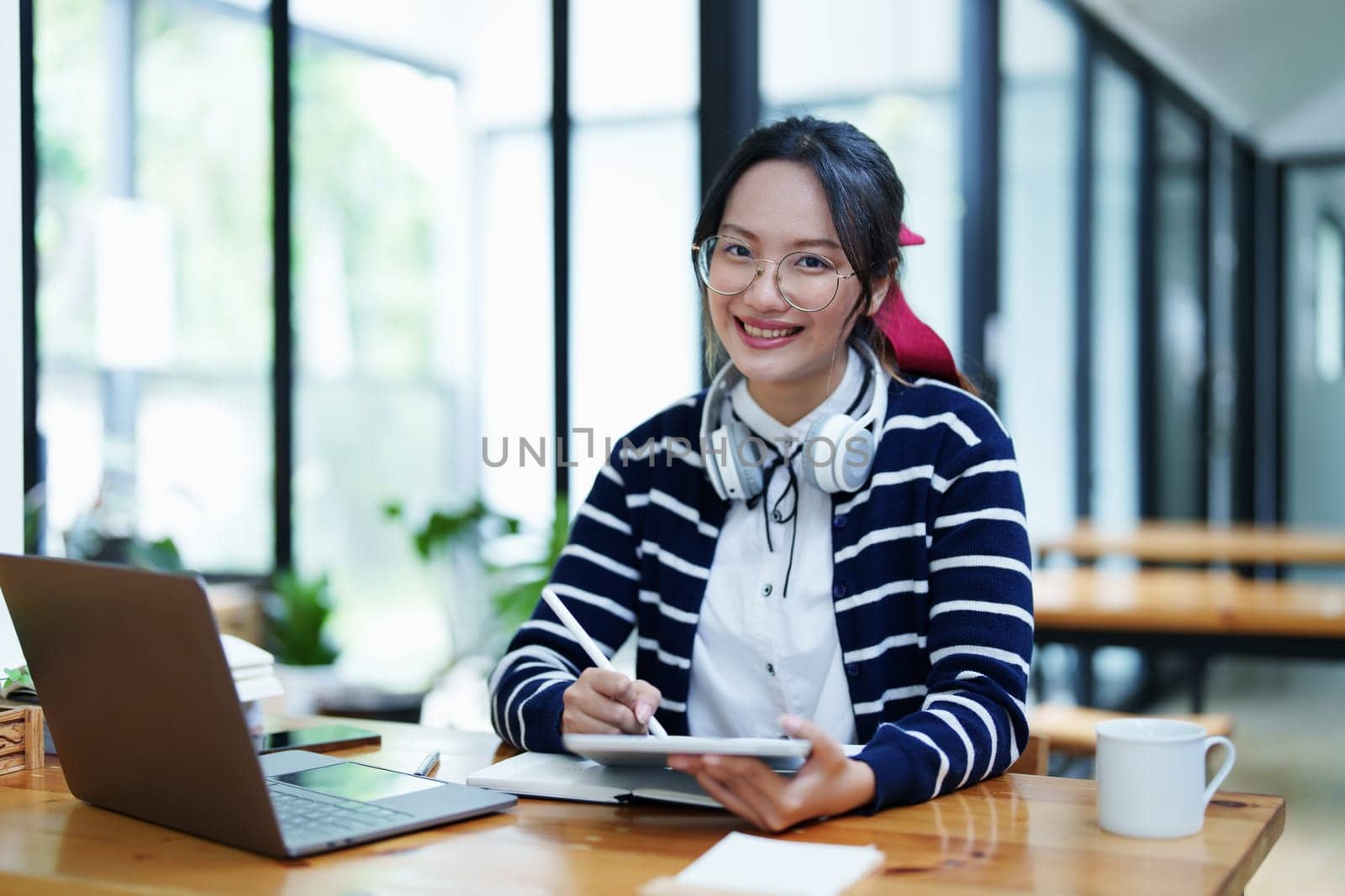 Portrait of a teenage Asian woman using a tablet, wearing headphones and using a notebook to study online via video conferencing on a wooden desk in library by Manastrong