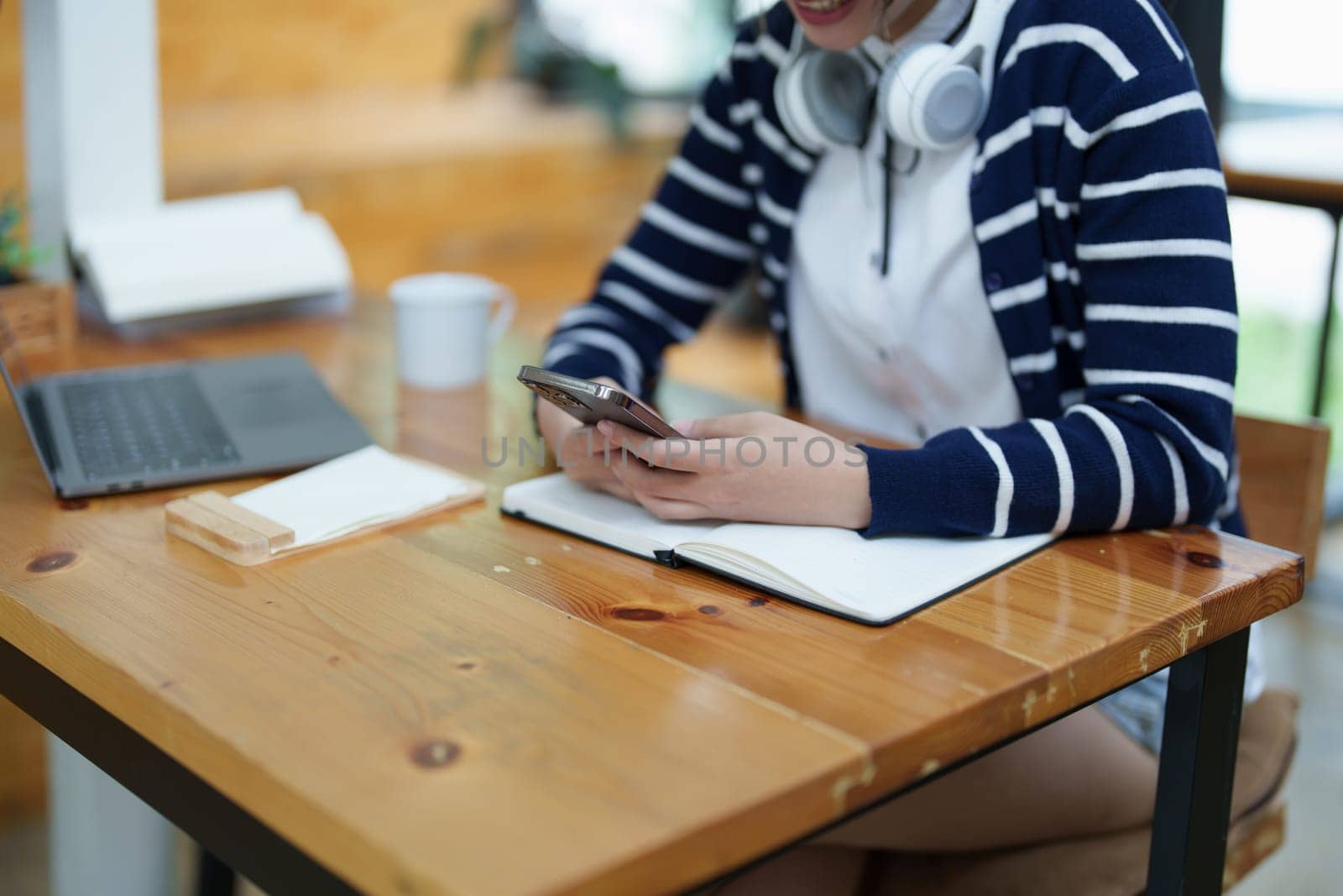 Portrait of a teenage Asian woman using a smartphone, wearing headphones and using a notebook to study online via video conferencing on a wooden desk in library.