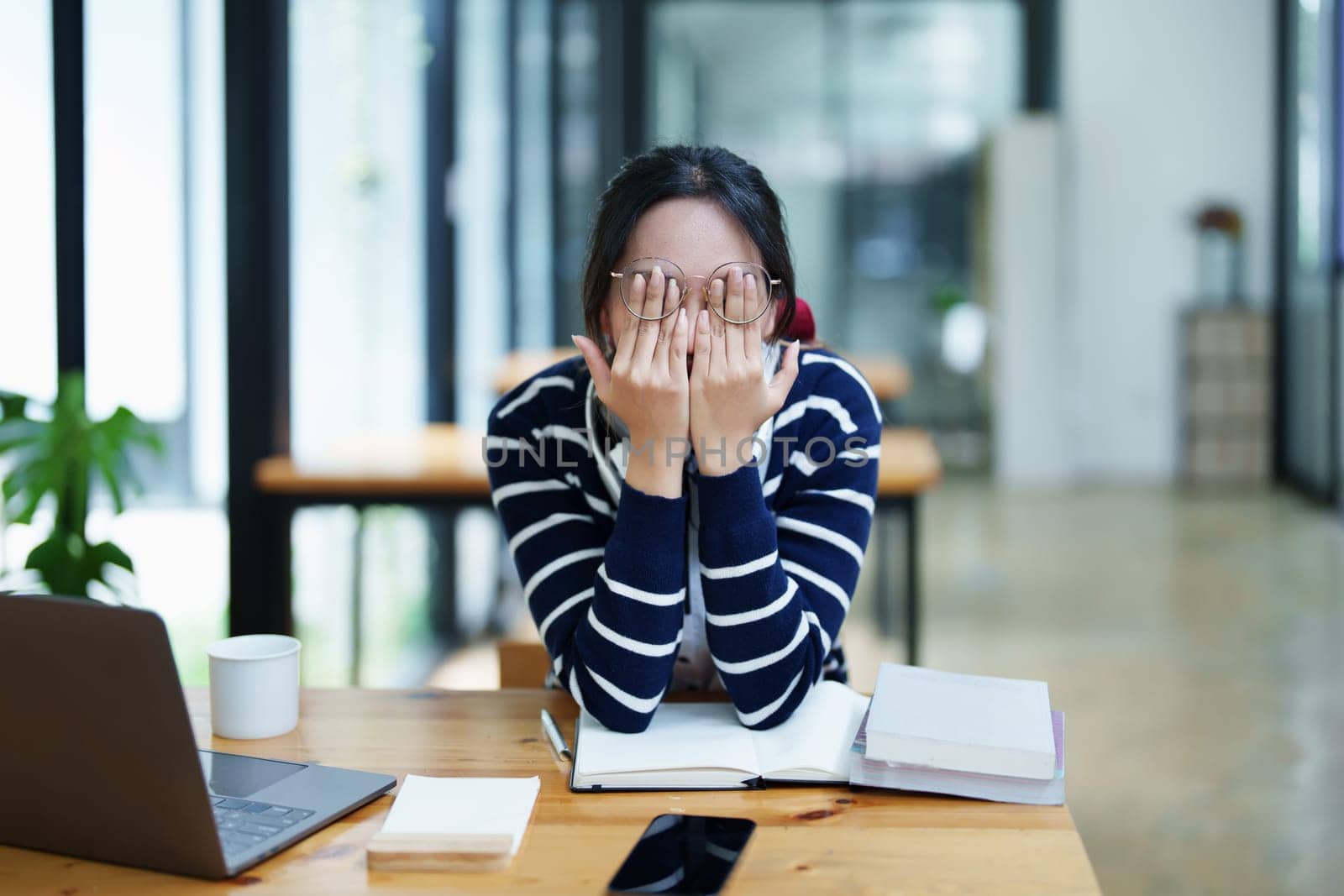 A portrait of a young Asian woman using a computer, wearing headphones and using a notebook to study online shows boredom and pain from video conferencing on a wooden desk in library.