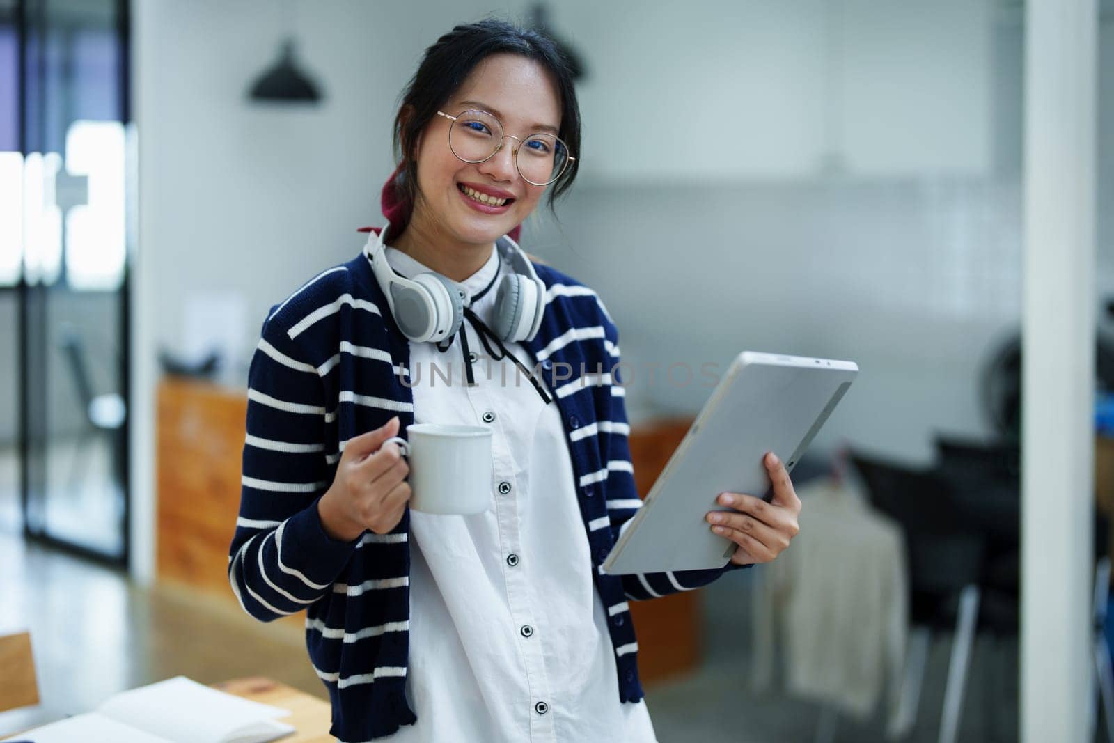 Portrait of a teenage Asian woman using a tablet, wearing headphones and using a notebook to study online via video conferencing on a wooden desk in library.