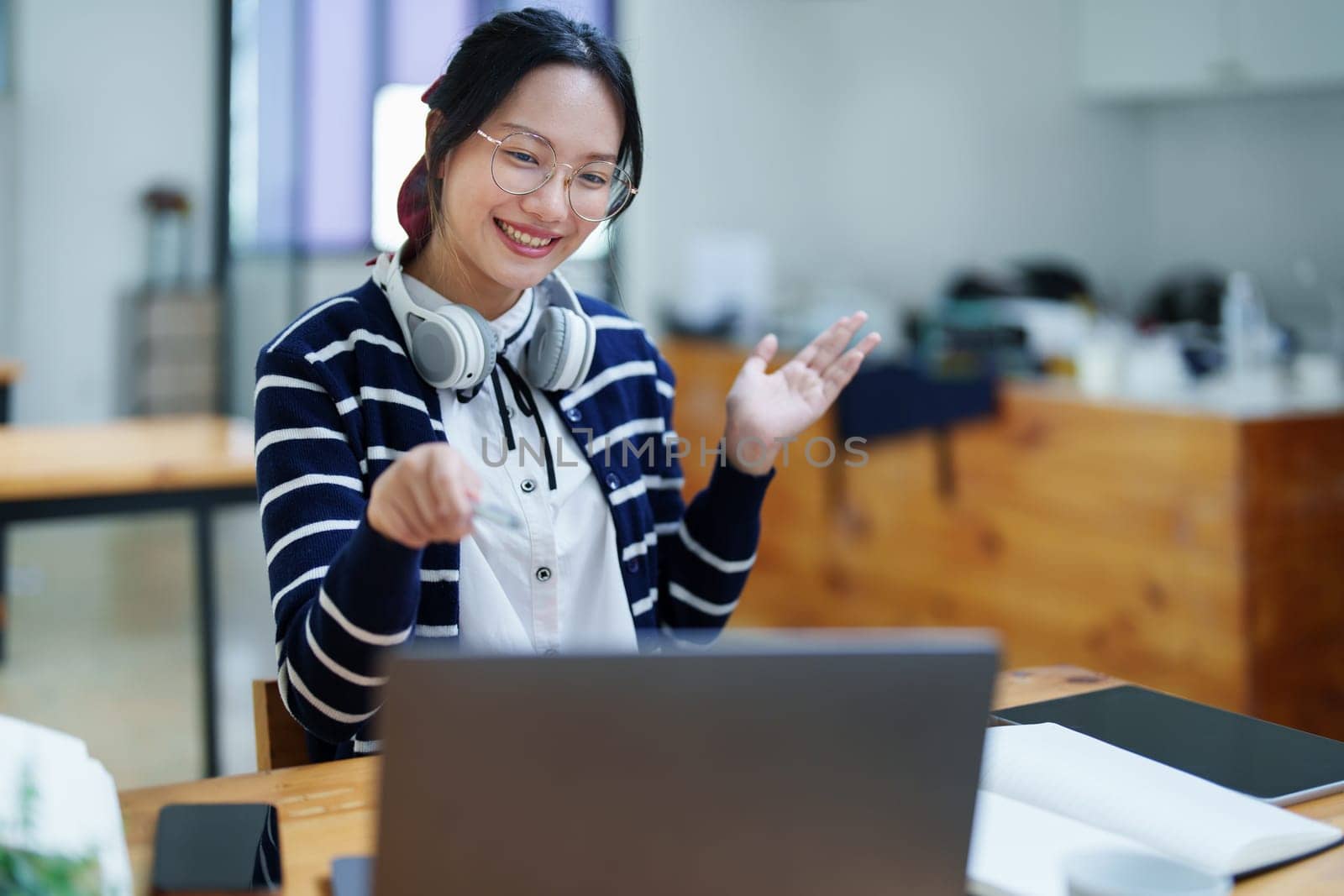 Portrait of a teenage Asian woman using a computer, wearing headphones and using a notebook to study online via video conferencing on a wooden desk in library.