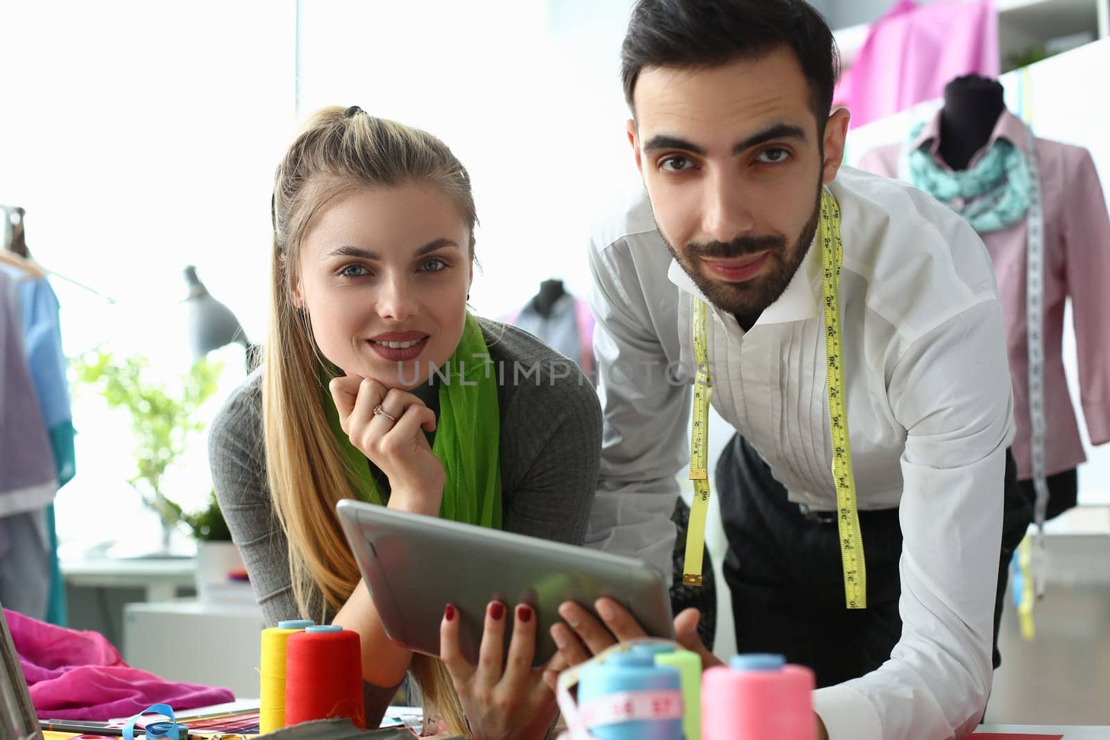 Man and woman designers are discussing new collection of clothes on digital tablet in workshop. Workers of online fashion store in modern warehouse.