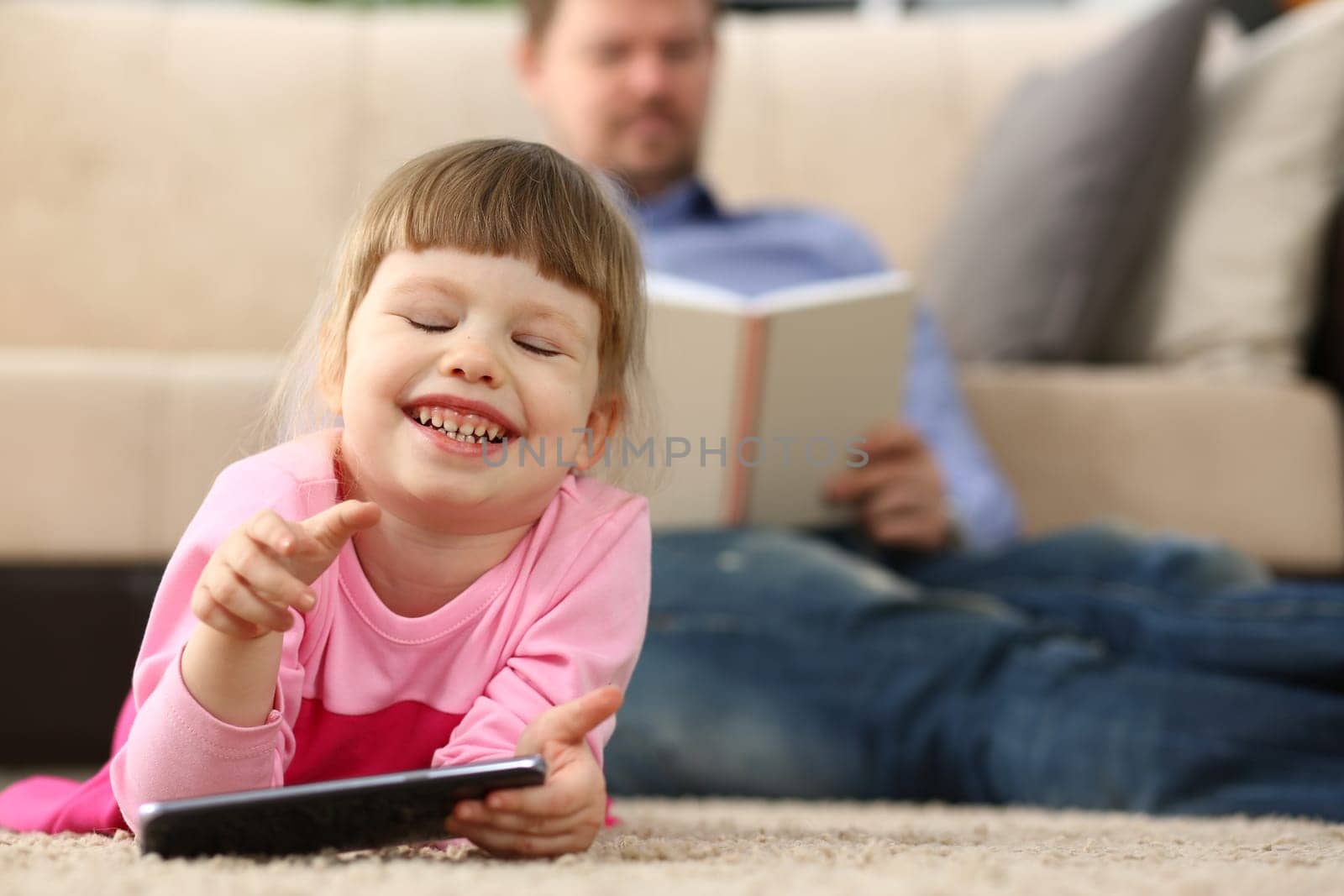 Little smiling baby girl with smartphone lies on floor in background dad reads a book by kuprevich