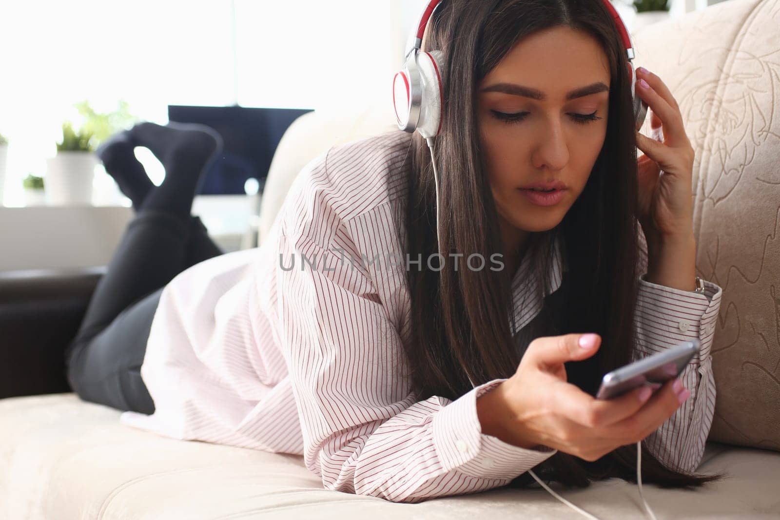 Young woman in headphones is relaxing on sofa and listening to music online using smartphone by kuprevich