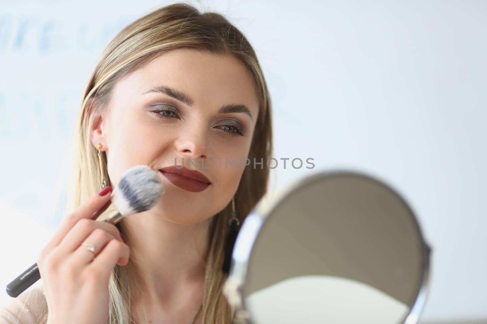 Smiling young woman looks in the mirror and paints herself with blush brushes. Makeup artist girl doing makeup learning with beauty and cosmetics video tutorials