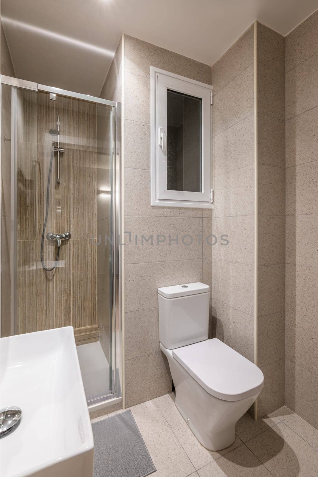 Multifunctional small bathroom with stylish beige tiles, glass shower door, washbasin, toilet and window. Concept of modern bathroom in a hotel. Single persons apartment by apavlin