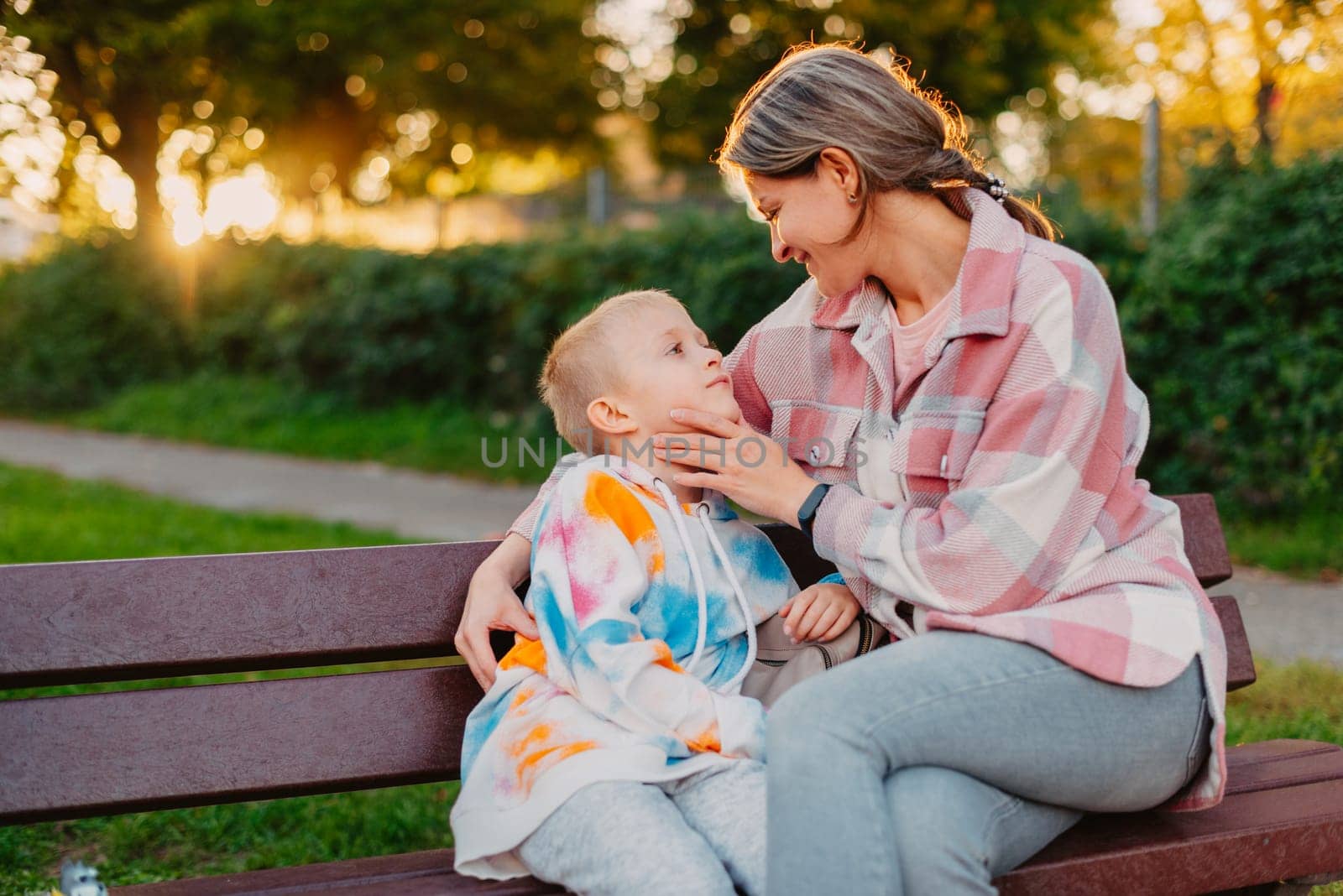 mother and son sit on a park bench in the rays of the setting sun. the concept of a family. Mother's Day. beautiful girl (mother) with a boy (son) in the park in the park are sitting on a bench at sunset.