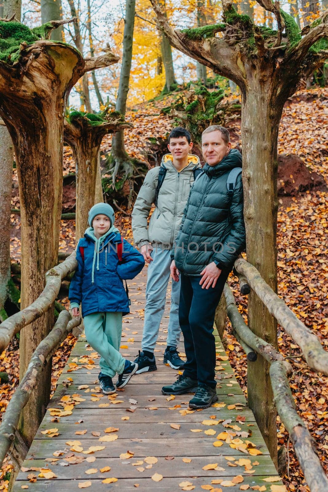 dad and 2 sons in the autumn forest stand on the bridge. Happy family enjoy the spending vacation together in the nature. father and son hug in the wooden pier at the background river and fall leaves forest by Andrii_Ko