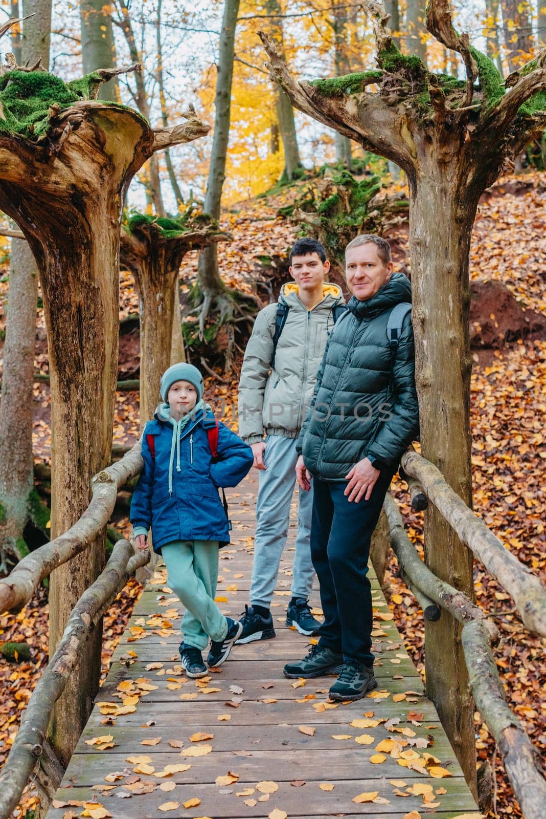 dad and 2 sons in the autumn forest stand on the bridge. Happy family enjoy the spending vacation together in the nature. father and son hug in the wooden pier at the background river and fall leaves forest by Andrii_Ko