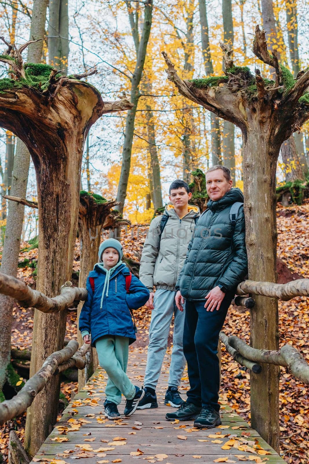 dad and 2 sons in the autumn forest stand on the bridge. Happy family enjoy the spending vacation together in the nature with favorite dog. Mother, father and son hug in the wooden pier at the background river and fall leaves forest