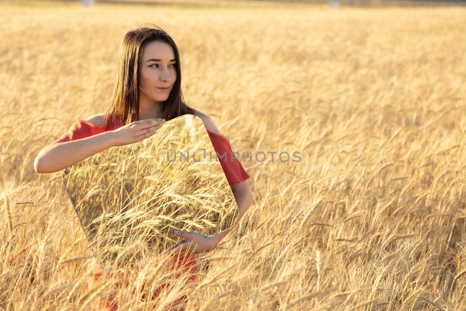 Smiling Young woman in the wheat field, holding mirror glass where reflected the dry wheat. Meditation, mental health concept. Copy space.