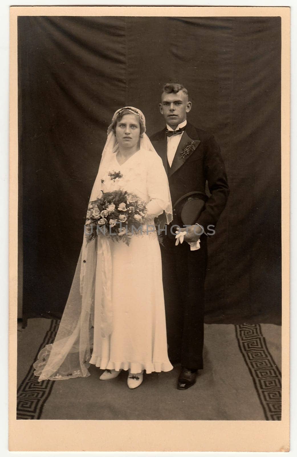 Vintage photo of newlyweds. Bride wears a long veil and holds wedding bouquet. Groom wears black suit and bow tie. Black white antique studio portrait. by roman_nerud