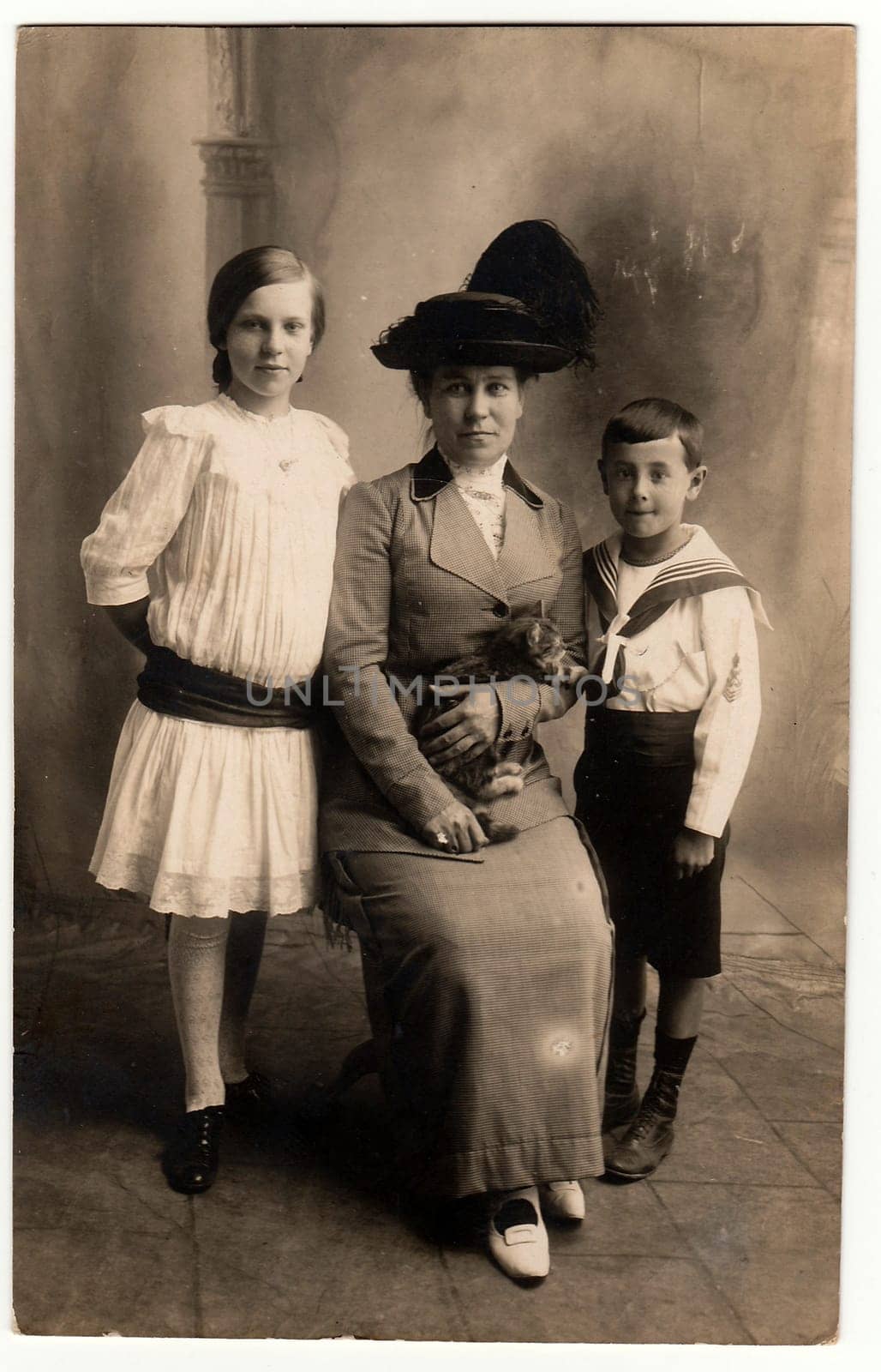 Vintage photo shows mother with her children girl and boy . Mother wears an elegant ladies costum and feather hat, she holds the cat. Boy wears sailor costum and girl wears white dress with black sash. by roman_nerud