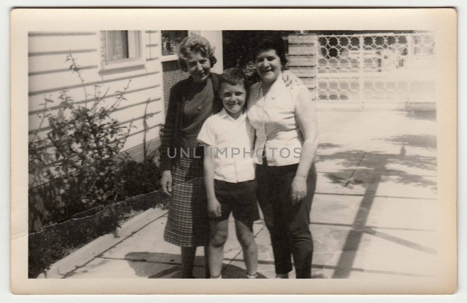 Retro photo shows women and boy pose in front of house. by roman_nerud