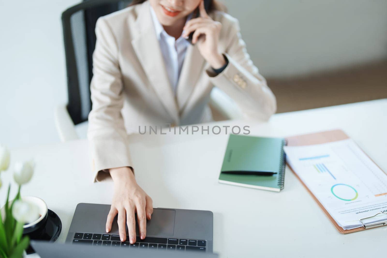 Portrait of a young Asian woman showing a smiling face as she uses her phone, computer and financial documents on her desk in the early morning hours by Manastrong