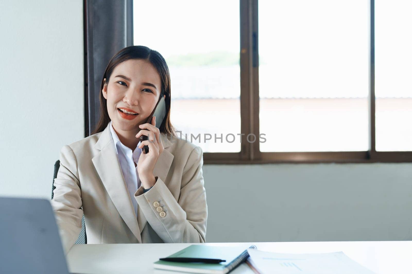 Portrait of a young Asian woman showing a smiling face as she uses her phone, computer and financial documents on her desk in the early morning hours.