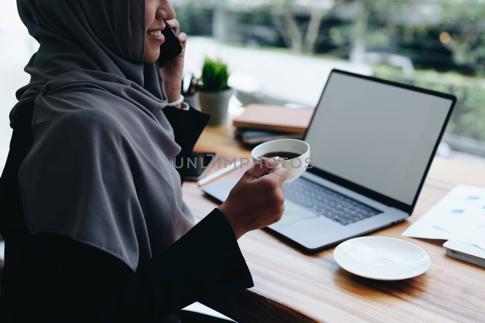 Beautiful Muslim woman talking on the phone and using computer on top while having coffee while working by Manastrong