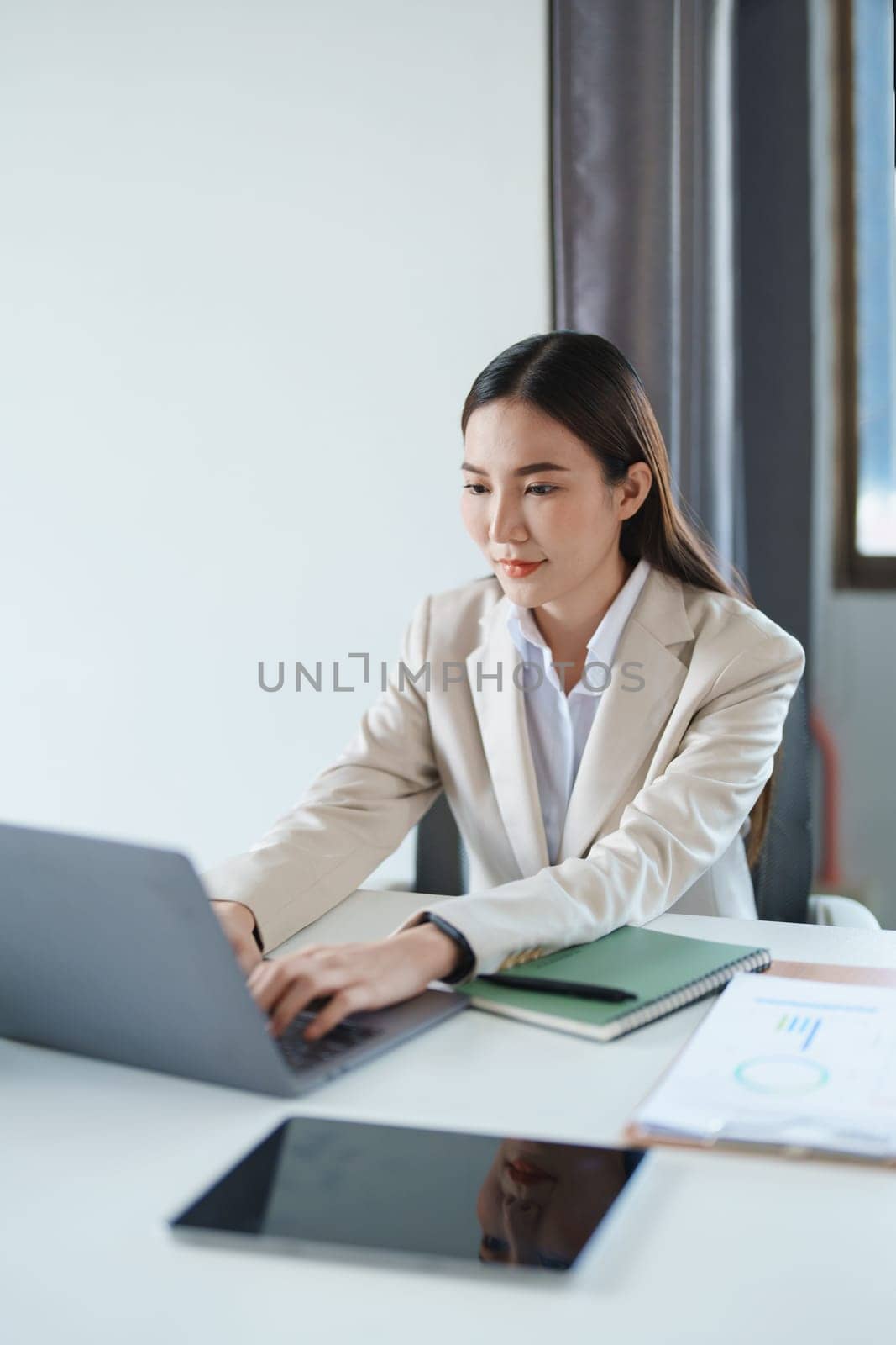 Portrait of a young Asian woman showing a smiling face as she using computer and financial documents on her desk in the early morning hours. by Manastrong