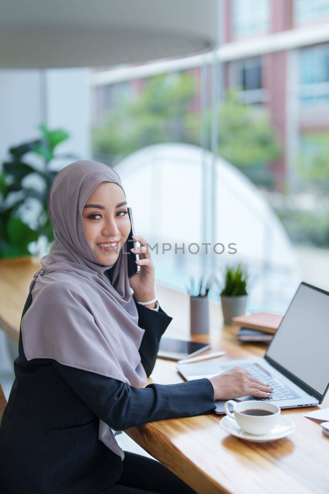 Beautiful Muslim woman talking on the phone and using computer on top while having coffee while working.