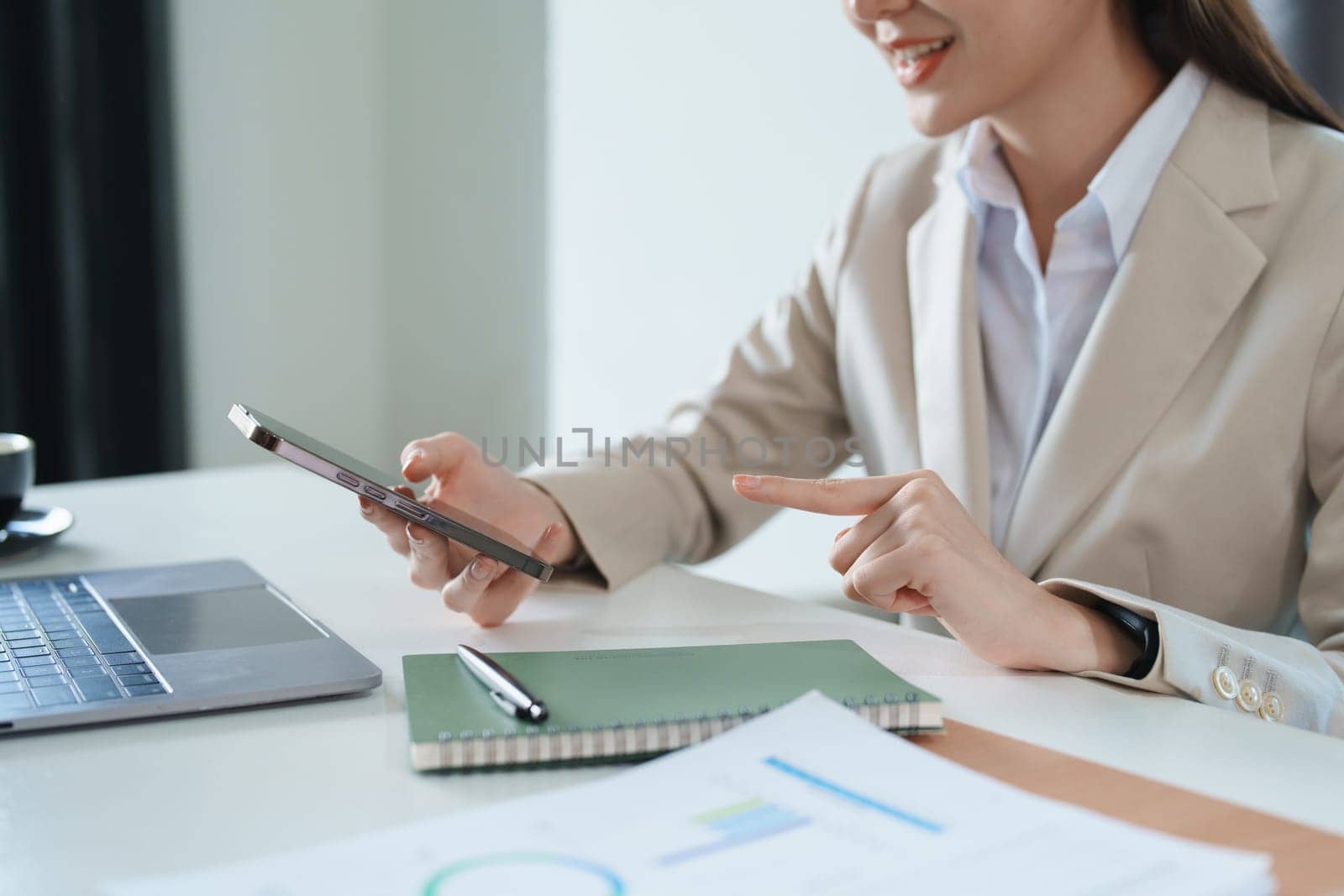 Portrait of a young Asian woman showing a smiling face as she uses her phone, computer and financial documents on her desk in the early morning hours by Manastrong