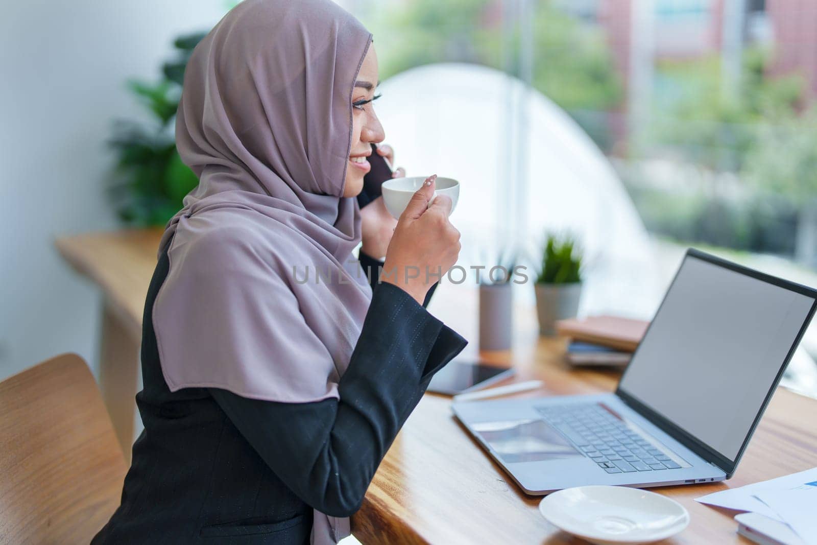 Beautiful Muslim woman talking on the phone and using computer on top while having coffee while working by Manastrong