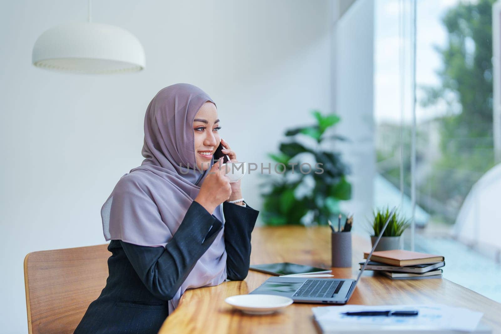 Beautiful Muslim woman talking on the phone and using computer on top while having coffee while working.