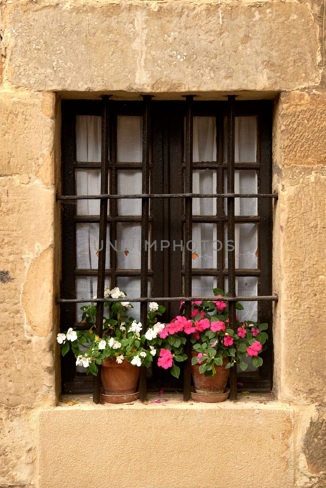 A window of a rustic house with railings and flowerpots,Coloured flowers, wrought iron, white curtains, stone, etc.